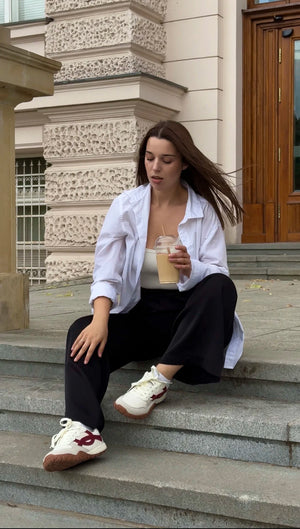 A woman sits on stone steps outside a building, wearing a white button-up shirt, black pants, and white sneakers with red accents while holding an iced coffee.