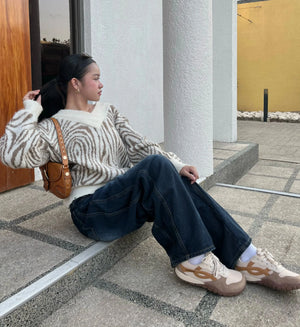 A woman sits on concrete steps wearing a cream and brown zebra-patterned knit sweater, dark wide-leg jeans, beige and white sneakers, and carries a brown leather shoulder bag.