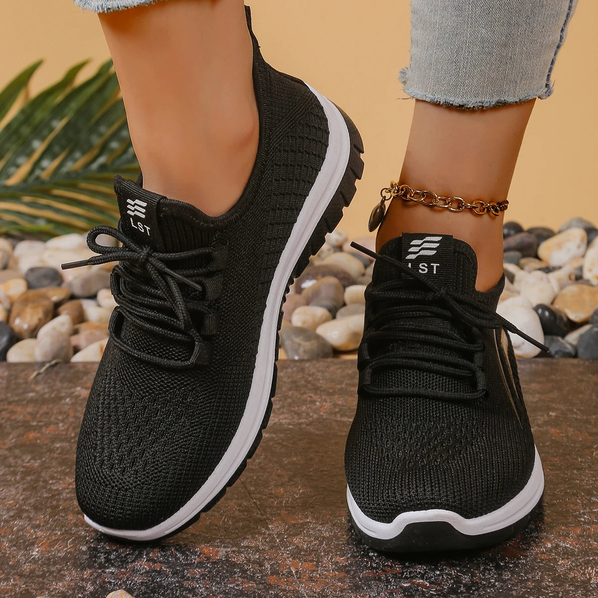 Black sneakers with white soles worn by a person, on a natural stone surface with pebbles and plants.