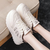 A pair of chunky low-top sneakers in off-white and beige, shown being worn with the feet resting on a beige upholstered bench. The shoes feature thick platform soles, lace-up fronts, and decorative side stripes.